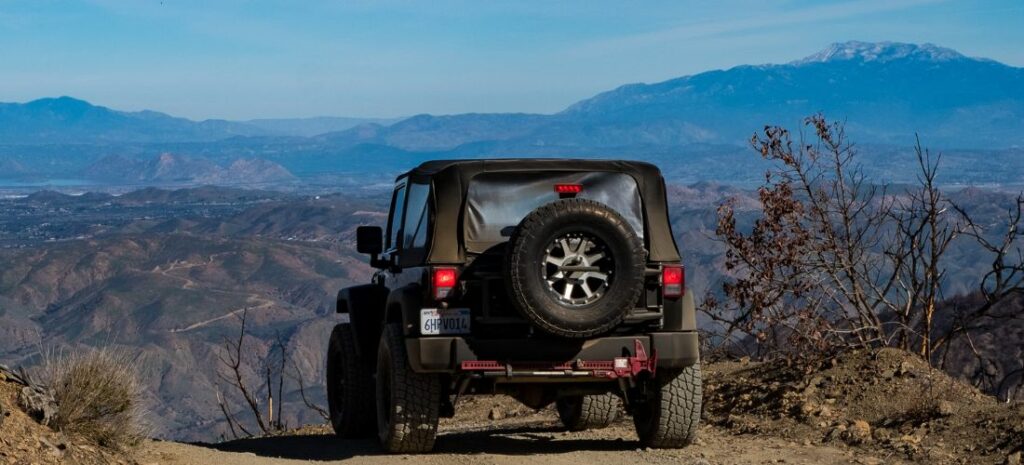 Stunning Jeep Wrangler with soft black top glistening in the sunlight, set against a spectacular mountain panorama under a clear blue sky.