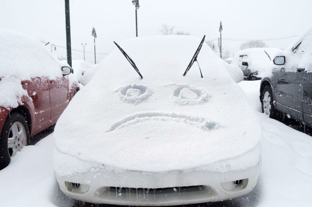 Winter storm car covered with snow with a sad face expression drawn on the hood of the car.