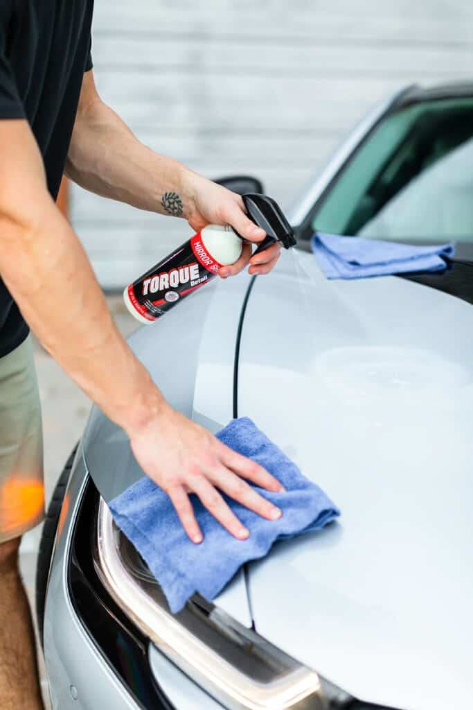 A man spraying Torque Detail Mirror Shine to his silver car while holding a blue microfiber towel in his right hand.